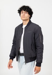 Zwarte bomberjack met geribbelde manchetten en zoom, zijzakken en een borstzak. Soepele stof met een subtiel logo detail aan de voorkant.