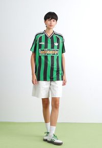 Žalias ir juodas dryžuotas sportinis marškinėlis su V formos iškirpte, ant kurio yra logotipas. Pora su baltais dryžuotais šortais ir žaliais sportbačiais.