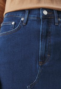 Jeans de denim azul oscuro en una persona que lleva un suéter beige, mostrando el bolsillo frontal, el botón, las presillas del cinturón y los detalles de costura.