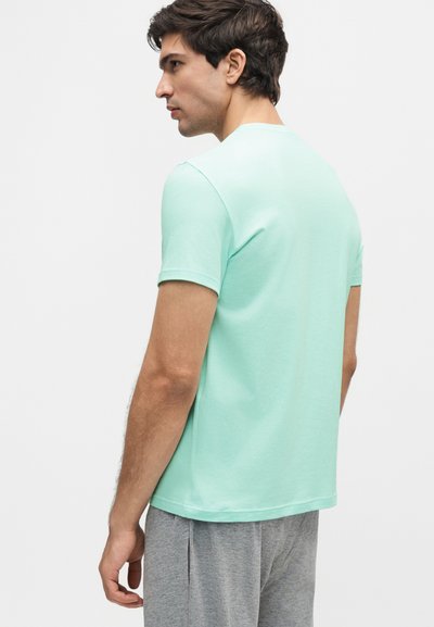 Νέος άνδρας που κοιτάζει προς τα αριστερά φορώντας ένα ανοιχτό μπλε κοντομάνικο μπλουζάκι και γκρι παντελόνι μπροστά σε ένα απλό λευκό φόντο.