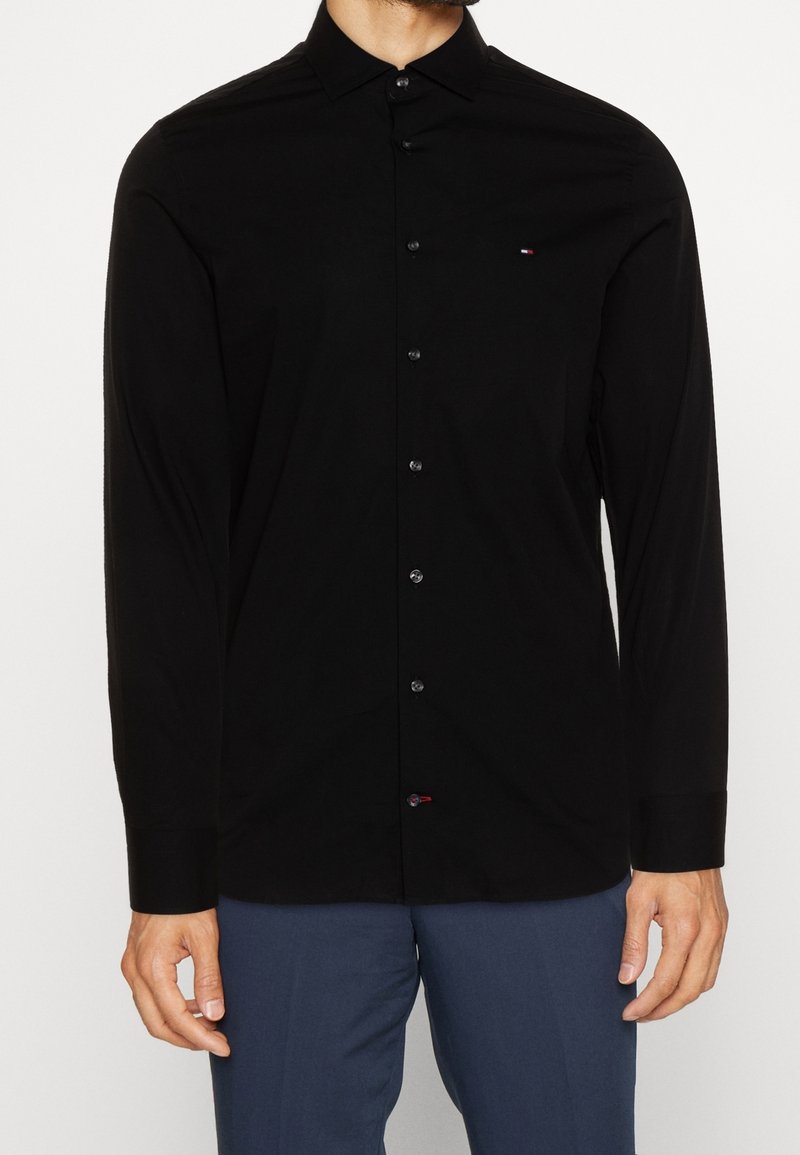 Camisa negra de manga larga hecha de tejido suave, con cierre de botones en la parte delantera, cuello puntiagudo y un pequeño logotipo en el lado izquierdo del pecho.