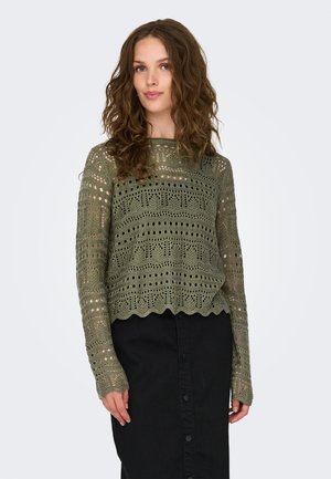 Pull en tricot vert olive avec des motifs ajourés, manches longues, ourlet festonné, associé à une jupe noire à boutons.