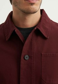 Veste chemise bordeaux avec un col, poche sans couture et accents en bouton noir ; fabriquée en tissu texturé, montrant une construction robuste.
