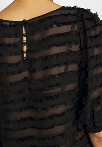 Svart topp med genomskinligt tyg, med horisontella strukturerade ränder. Detalj med nyckelhål i ryggen med knappstängning och fransade kanter på ränderna.