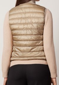 Nainen, joka on pukeutunut beigeen tikkivestiin vaaleanpunaisen pitkähihaisen neuleen ja mustien housujen päälle, nähtynä takaa.