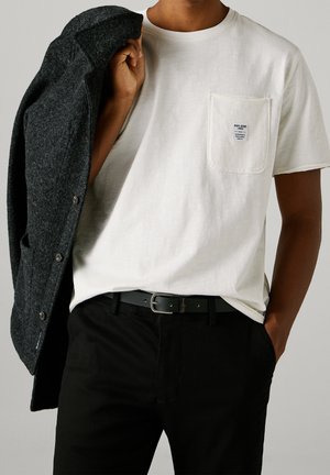 Mann trägt weißes T-Shirt mit Taschenetikett, schwarze Hose und schwarzen Gürtel, hält eine dunkelgraue Jacke über der Schulter vor neutralem Hintergrund.