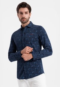Marineblauwe lange mouwen shirt met bloemenpatroon, knoopsfeer aan de voorkant en kraag. Kenmerkt een rode logoaccent op de linkerkant van de borst. Soepele textuur.