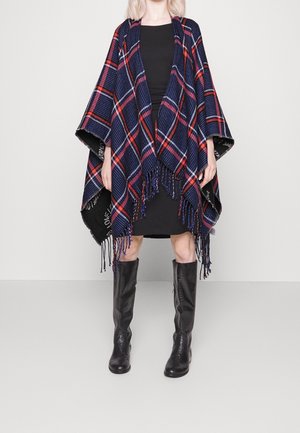 Kobieta ubrana w niebiesko-czerwoną kratkowaną ponczę z frędzlami na czarnej sukience, zestawioną z czarnymi, skórzanymi kozakami za kolano, stojąca na białej podłodze.