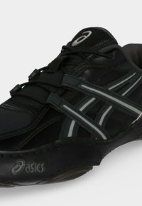 Μαύρο αθλητικό παπούτσι Asics με γκρι και λευκές ρίγες, ελαστικά κορδόνια και υφή στη σόλα.