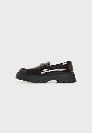 Loafer maro închis lucios, cu talpă neagră groasă și emblemă decorativă pe cataramă, văzut din lateral pe un fundal simplu.