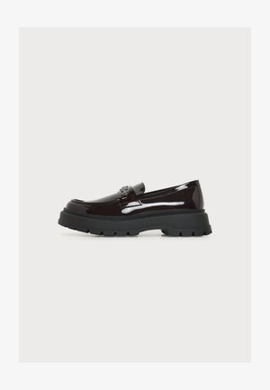 Glänzender dunkelbrauner Loafer mit klobiger schwarzer Sohle und dekorativem Emblem auf dem Riemen, Seitenansicht vor schlichtem Hintergrund.