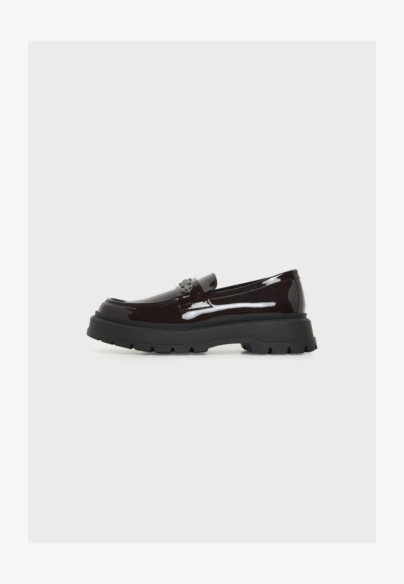 Mocassin marron foncé brillant avec semelle noire épaisse et emblème décoratif sur la bride, vu de profil sur un fond uni.