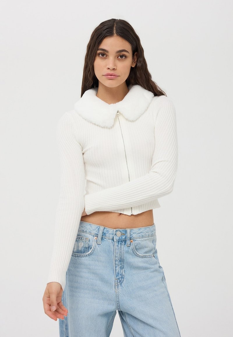 Biała prążkowana krótka sweter z kołnierzem z sztucznego futra i zapięciem na zamek. Połączony z jasnoniebieskimi dżinsami o wysokim stanie.
