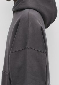 Sweat à capuche gris foncé en tissu doux et texturé. Comprend une grande capuche et des coutures latérales avec surpiqûres contrastées. Poche avant visible.