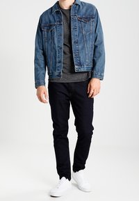 Tmavě modrá džínová bunda, pod ní šedý svetr, černé džíny a bílé tenisky. Bunda má kapsy a zapínání na knoflíky.