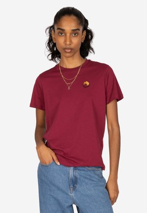 Frau mit dunklen Haaren, die ein bordeauxfarbenes T-Shirt mit einem kleinen Croissant-Design und blaue Jeans trägt, posiert mit einer Hand in der Tasche.