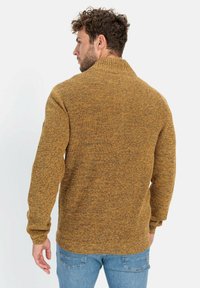 Senfgelbes Strickpullover mit strukturiertem Finish, featuring einen hohen Kragen und gerippte Bündchen. Getragen mit hellblauen Jeans.