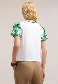 Top bianco con maniche corte a volant verdi a fantasia, caratterizzato da una vestibilità comoda. Pantaloni marroni parzialmente visibili, con un braccialetto a maniche rotonde al polso.