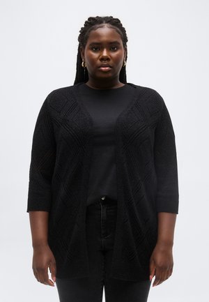 Čierna žena s vrkočmi, oblečená v čiernom štruktúrovanom kardigáne, čiernej košeli, čiernych nohaviciach a zlatých kruhových náušniciach, stojaca pred obyčajným pozadím.