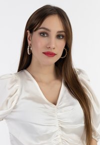 Bluză albă din satin cu mâneci bufante și decolteu în V, având detalii pliate în față. Cercei mari aurii cu perle completează look-ul.