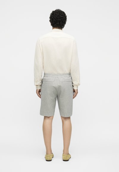 Άνδρας με σγουρά μαλλιά, που φοράει κρεμ μακρυμάνικο πουκάμισο, γκρι σορτς μέχρι το γόνατο και μπεζ παπούτσια χωρίς κορδόνια, στεκόμενος με την πλάτη προς το μέρος μας.