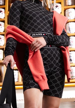 Osoba w czarnym stroju Tommy Jeans z wzorem uśmiechniętej buźki, trzymająca koralową bluzę, stojąca przed drewnianymi półkami z słoikami.