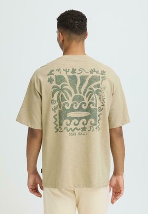 Homme aux cheveux courts et bouclés portant un t-shirt beige avec un motif tropical vert de palmiers, vagues, planche de surf, et le texte "RESTEZ SALE" au dos.