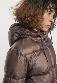 Person med krøllet hår iført en skinnende brun dunjakke med hætte og sorte snore, set fra siden mod en ensfarvet baggrund.