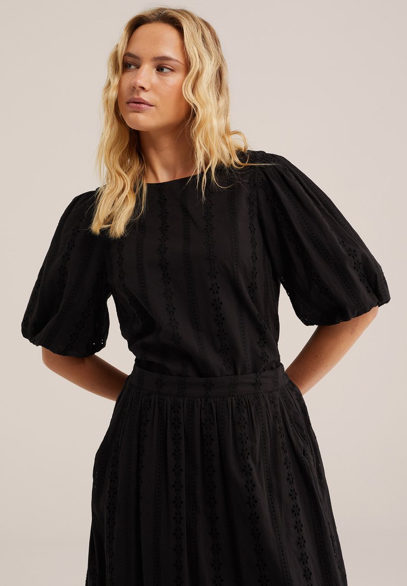 Mujer rubia con cabello ondulado que lleva un vestido negro con mangas cortas abullonadas y sutil bordado floral, posando contra un fondo liso.