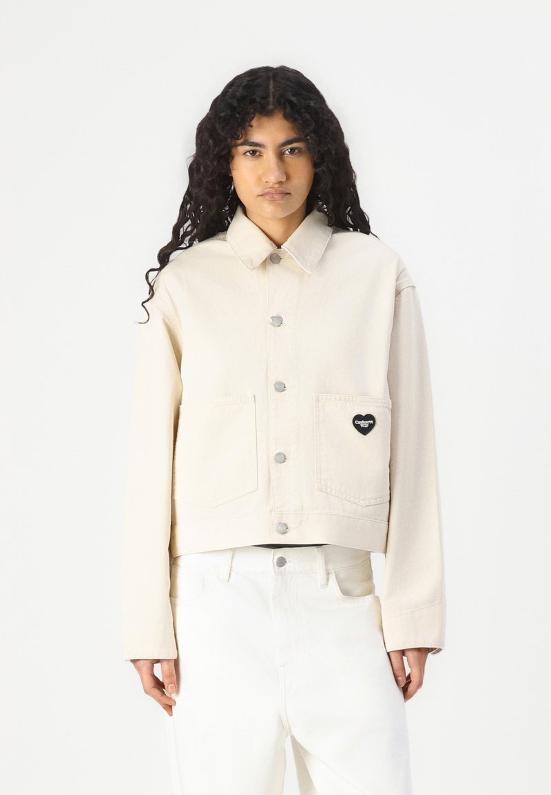 Cream-colored cropped jacket made of denim with a button front, two front pockets, and a heart-shaped logo patch on the left pocket.