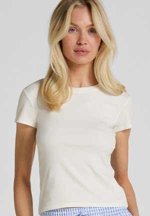 Blonde vrouw met schouderlang haar, gekleed in een wit geribd T-shirt met korte mouwen en blauw gestreepte broek, kijkt recht vooruit tegen een effen achtergrond.