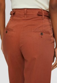 Pantalon couleur rouille avec une texture lisse, doté de passants de ceinture, de deux poches arrière à boutons, et d'une coupe ajustée.
