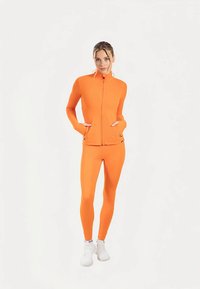 Survêtement de sport orange composé d'une veste zippée ajustée et d'un legging assorti. Tissu lisse et extensible avec manches longues et poches latérales.
