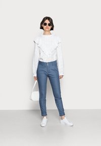 Hvit langermet bluse med volangkrage, kombinert med blå tettsittende bukser med knappedetaljer, og hvite sneakers. Liten hvit veske.
