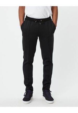 Man draagt zwarte slim-fit joggerbroek met tailleband met trekkoord, een wit t-shirt en zwarte sneakers met witte zolen, staat met handen in de zakken.