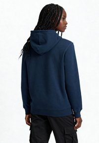 Hoodie bleu marine avec capuche à cordon de serrage, poignets et ourlet côtelés, texture douce et coupe décontractée. Porté avec un pantalon cargo noir.