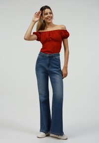 Red off-the-shoulder top with textured pattern, paired with high-waisted blue flared jeans and white sneakers. Model poses with hand raised.