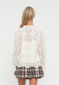 Vrouw met lang blond haar in een witte blouse van kant met bloemmotieven en een bruine geruite geplooide rok, staand met haar rug naar de kijker gericht.