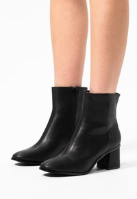 Bottines en cuir noir avec un bout pointu, un talon bloc de hauteur intermédiaire et une fermeture éclair sur le côté. Texture lisse avec des accents minimaux.