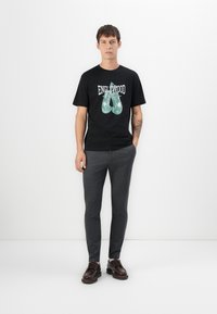Young man standing wearing black "Englewood" t-shirt with green boxing gloves print, gray pants, and brown leather shoes against white background.