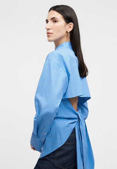 Hosszú sötét hajú nő, kék csíkos, hátul nyitott, masnival megkötött ingben, sötét farmerrel, balra néz semleges háttér előtt.