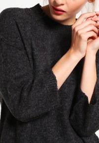 Pull en maille gris foncé avec une texture douce, une coupe ample et des manches larges, présentant un motif côtelé subtil au niveau de l'encolure et des poignets.