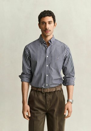 Man met kort krullend haar draagt een opgerolde gestreepte blouse met knopen, een bruine broek en een horloge, staand tegen een effen achtergrond.