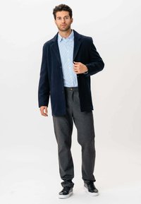 Ung mand står op, iført en marineblå blazer, lys blå stribet skjorte, mørkegrå bukser og sorte sneakers med hvide såler mod en hvid baggrund.