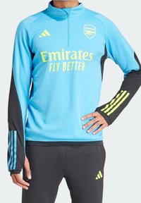 Ljusblå träningsöverdel med svarta ärmar, prydd med gul text "Emirates Fly Better" och Arsenal-logotyp, tillverkad av ett slätt material.