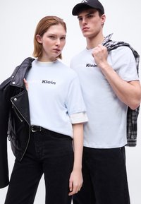 Νεαρή γυναίκα και άνδρας που φορούν ανοιχτά t-shirts Calvin Klein και μαύρα παντελόνια, κρατώντας ο καθένας ένα σακάκι στον ώμο τους μπροστά σε λευκό φόντο.