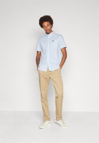Short-sleeve blue and white gingham shirt with a button-down collar and embroidered logo, paired with light beige trousers and white sneakers.