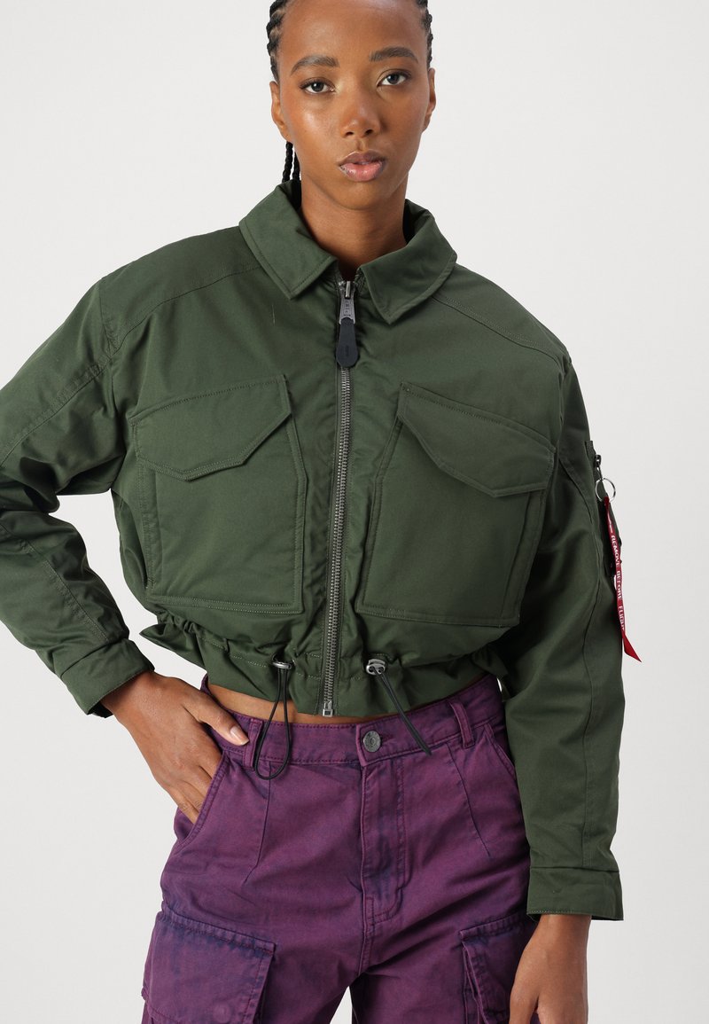 Mujer vestida con una chaqueta corta de color verde oscuro con cremallera y bolsillos con solapa, y pantalones cargo morados, de pie contra un fondo claro y liso.