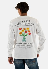 Camiseta blanca de manga larga con un gráfico de un jarrón de flores y texto negro que muestra el nombre del restaurante y detalles de ubicación en la parte posterior.