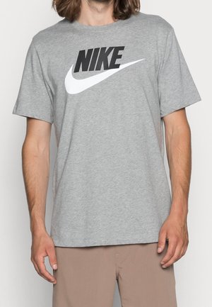Mężczyzna w szarej koszulce z czarnym napisem "NIKE" i białym logo swoosh, w połączeniu z jasnobrązowymi spodniami na jednolitym tle.
